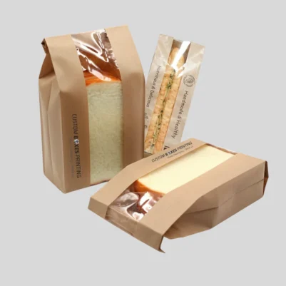 Two paper bags from a bakery, each sealed with plastic wrap, containing freshly baked bread.
